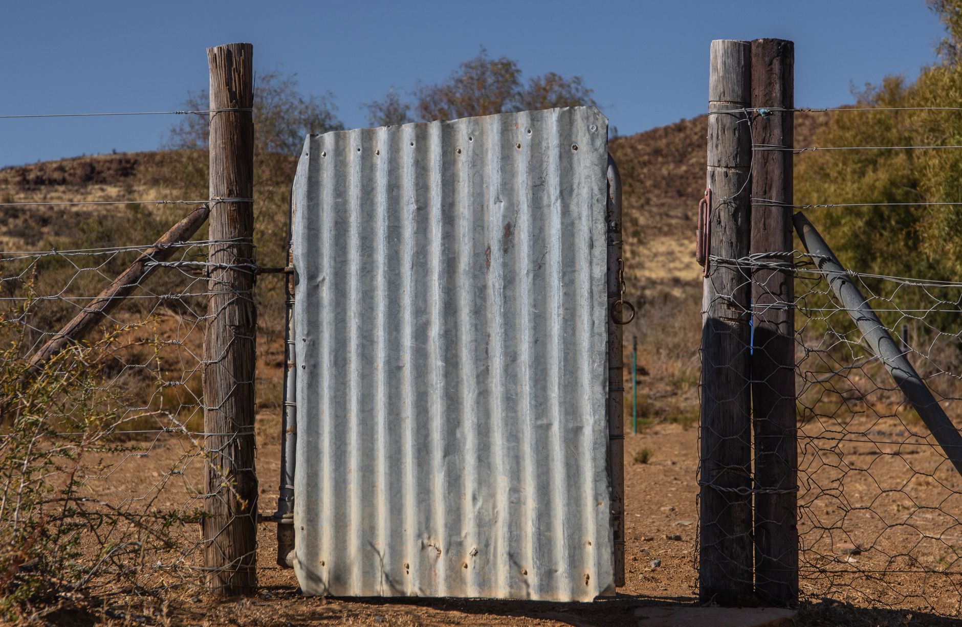 Corrugated metal gate framed by wooden posts in a dry, rugged landscape under clear skies